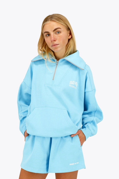 Osaka Women Half Zip Sweater | Blue Topaz Osaka women half zip sweater in light blue with white logo. Front flatlay view