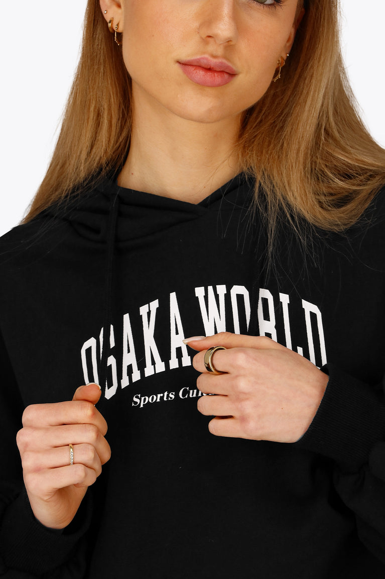 Woman wearing the Osaka women cropped hoodie in black with college logo in white. Front detail logo view