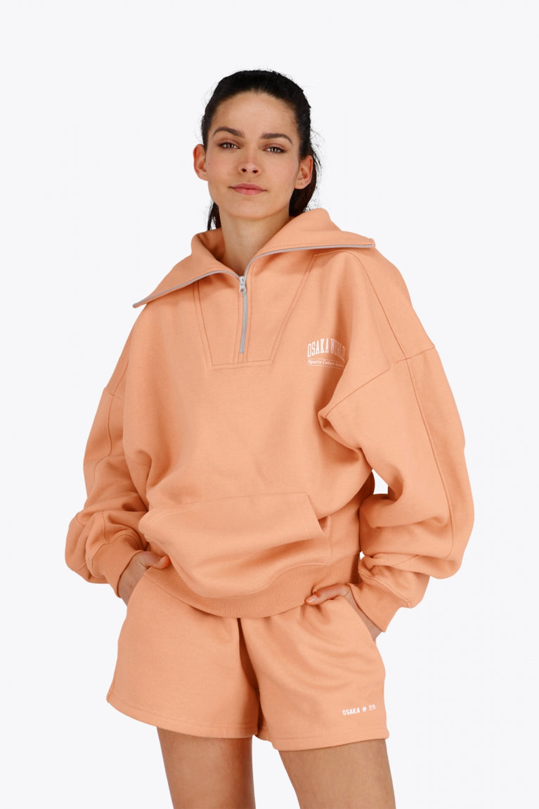 Woman wearing the Osaka women half zip sweater in peach with white logo. Front view