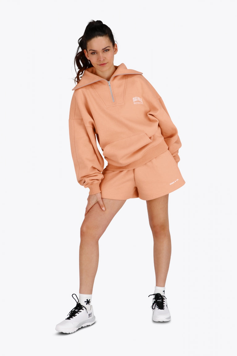 Woman wearing the Osaka women half zip sweater in peach with white logo. Front view