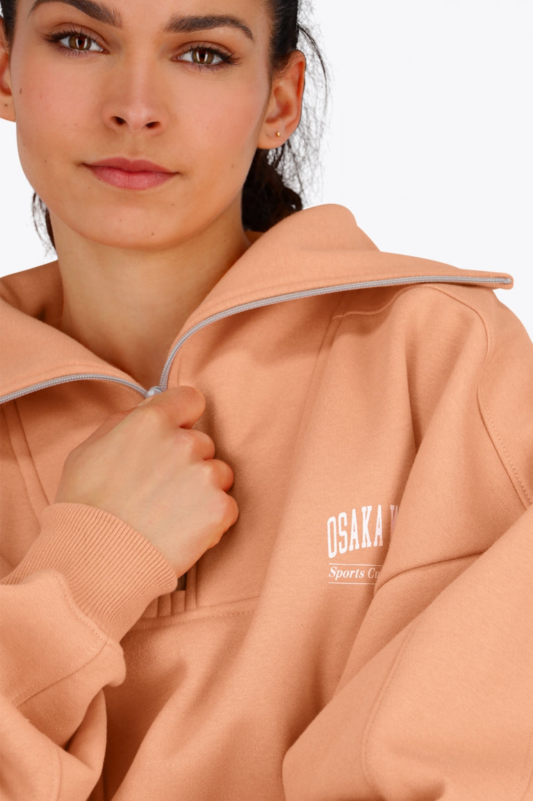 Woman wearing the Osaka women half zip sweater in peach with white logo. Front detail neck view