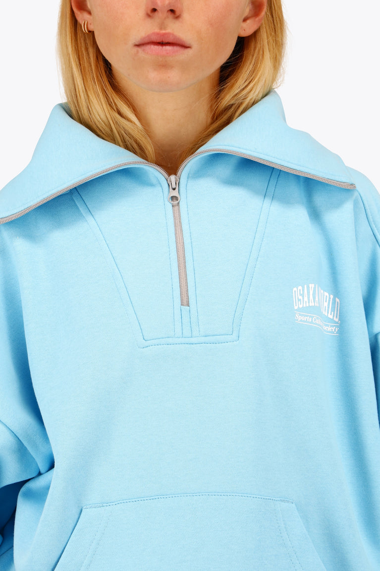 Woman wearing the Osaka women half zip sweater in light blue with white logo. Front detail neck view