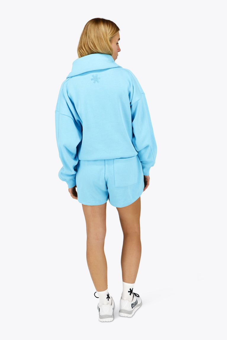 Woman wearing the Osaka women half zip sweater in light blue with white logo. Back view