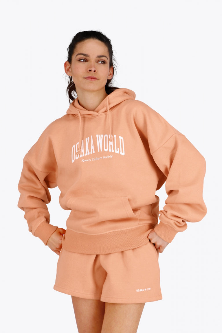 Woman wearing the Osaka women hoodie in peach with white logo. Front view