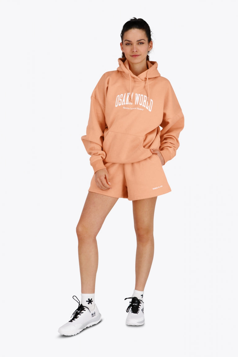 Woman wearing the Osaka women hoodie in peach with white logo. Front view