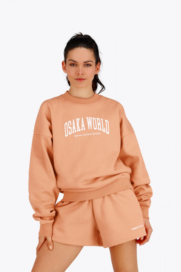 Woman wearing the Osaka women sweater in peach with logo in white. Front view
