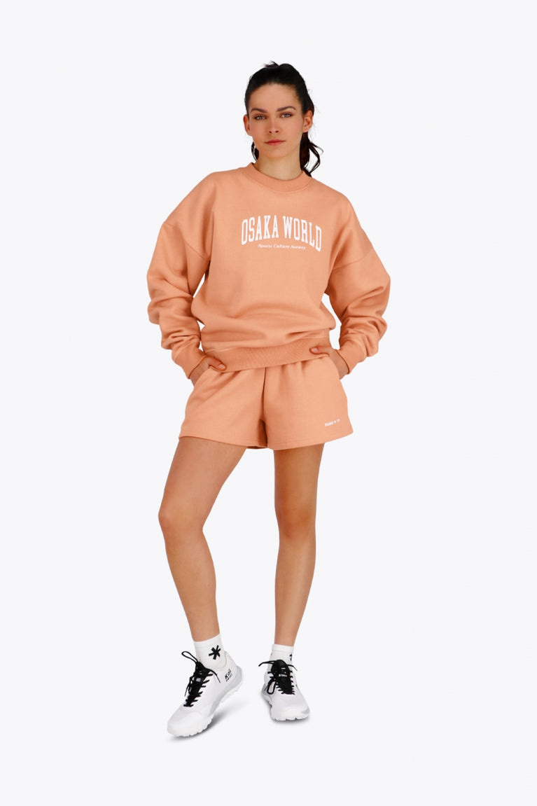 Woman wearing the Osaka women sweater in peach with logo in white. Front view