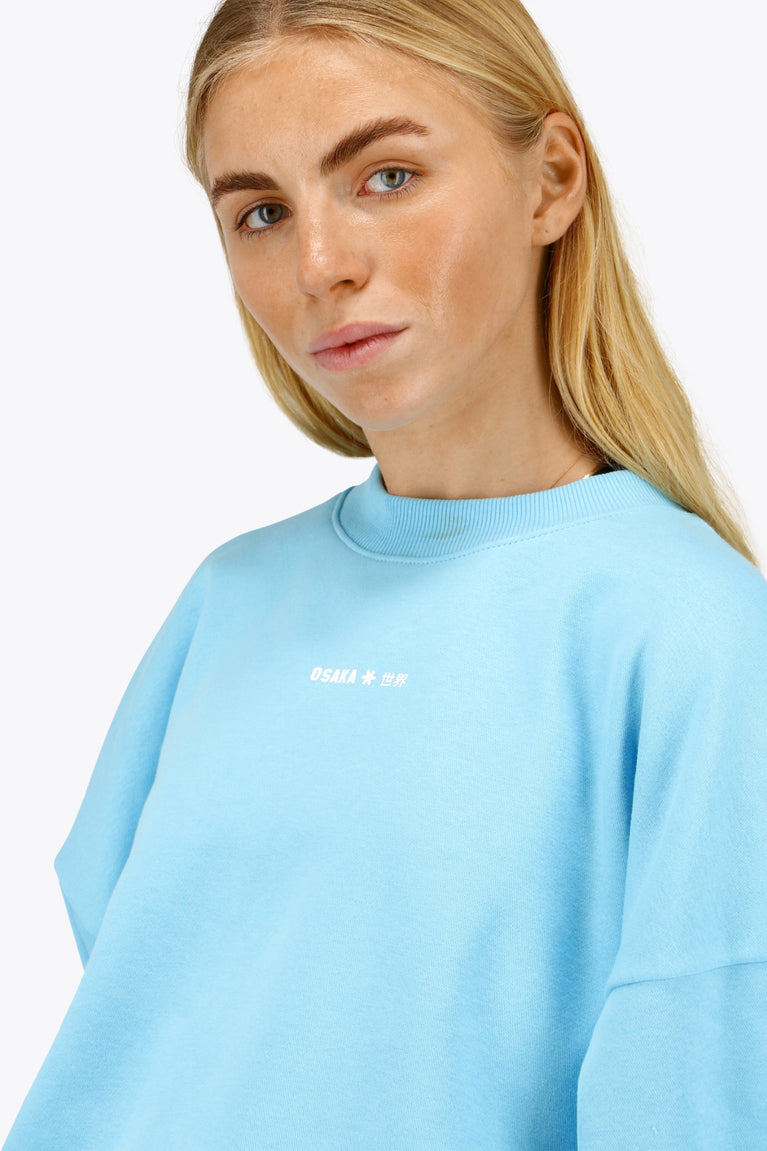 Woman wearing the Osaka women sweater in light blue with logo in white. Front view