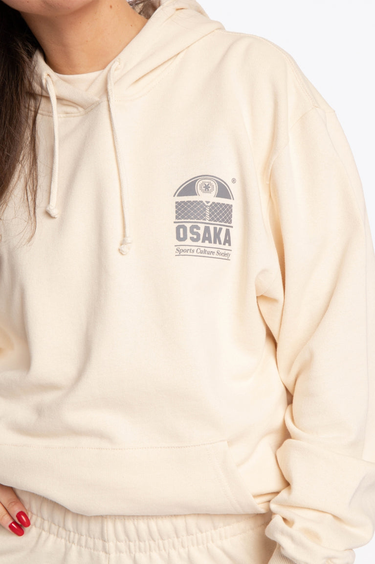 Woman wearing the Osaka unisex cream hoodie sports culture society with logo in dark grey. Front detail logo view