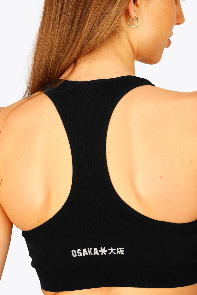 Woman wearing the Osaka women tech sports bra in black with logo in grey. Back detail view
