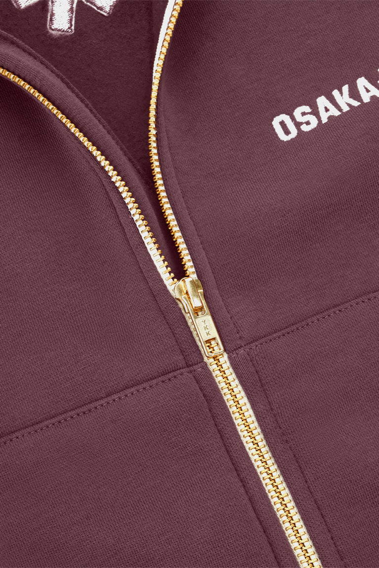 Plum zip-up sweater close-up showing gold zipper and embroidered chest logo