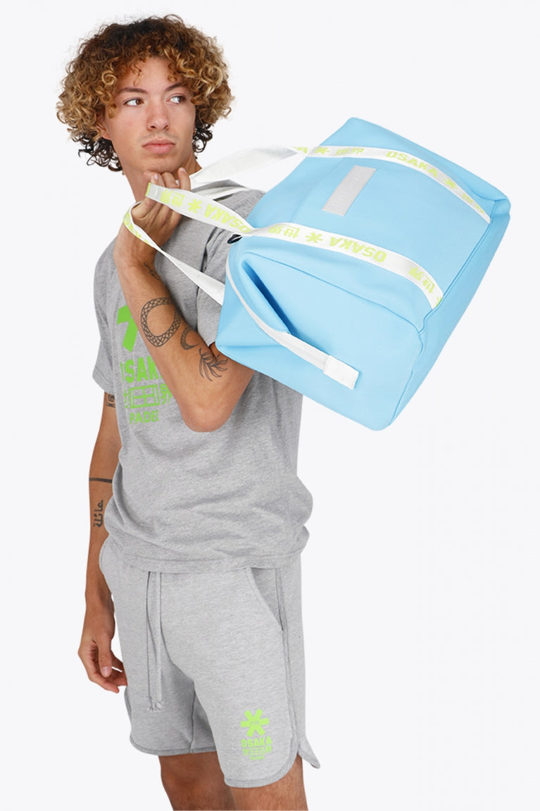 Osaka neoprene duffel bag in light blue with logo in white on the bag and in green on the straps. Man holding the bag