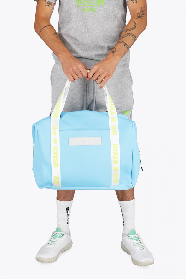 Osaka neoprene duffel bag in light blue with logo in white on the bag and in green on the straps. Man holding the bag