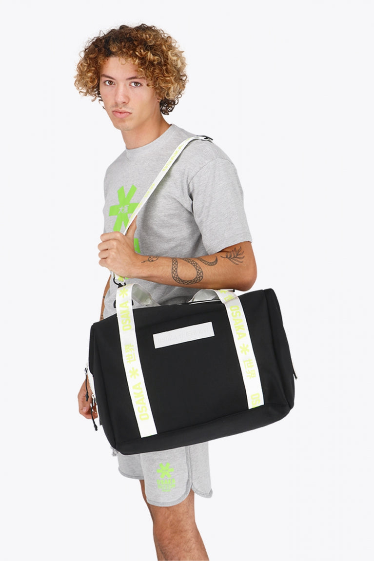 Osaka neoprene duffel bag in black with logo in white on the bag and in green on the straps. Man wearing the bag